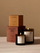 Two lit Olfacte scented candles, Chapter and Private View by Audo, in dark glass jars with their tan and terracotta boxes.