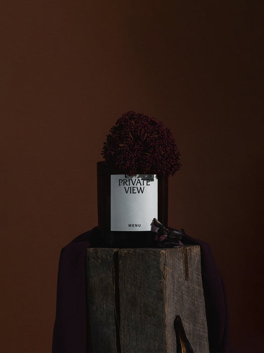 Audo's Olfacte Scented Candle in Private View, presented on a rustic wooden block with a dark burgundy flower on top.