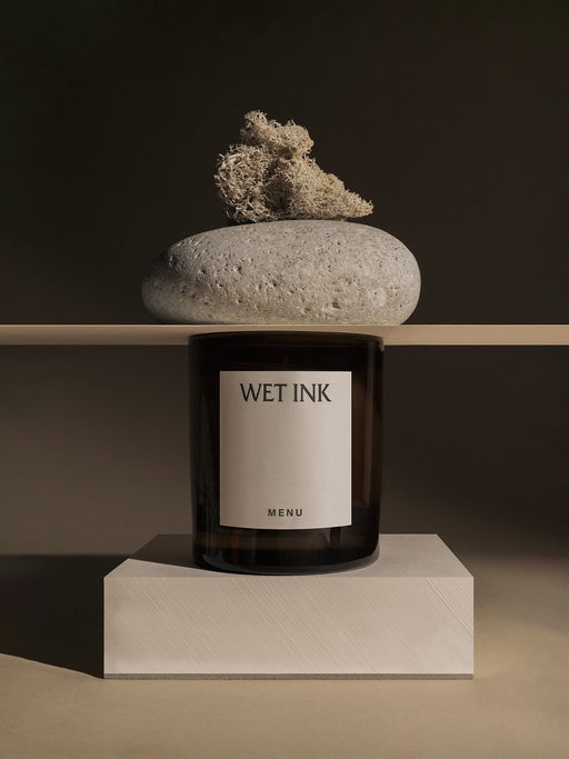 The Olfacte Scented Candle, Wet Ink, in its dark jar, is styled on a plinth below a shelf with a stone and dried moss.