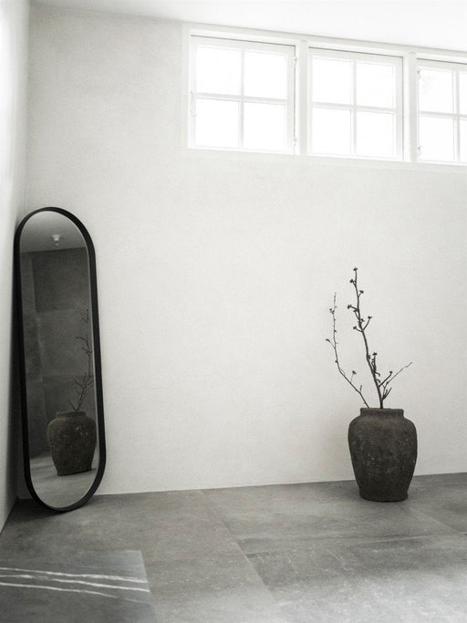 A black-framed Norm Oval Mirror leans in the corner of a minimalist room with grey floors, a stone vase, and high windows.
