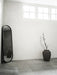 A black-framed Norm Oval Mirror leans in the corner of a minimalist room with grey floors, a stone vase, and high windows.
