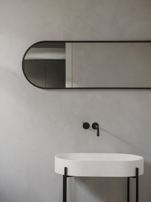 The black-framed Norm Oval Mirror mounted horizontally on a grey bathroom wall, above a white oval sink with a black tap.