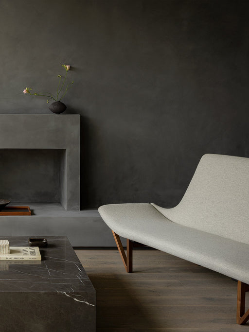 A light grey Pagode Sofa with wooden legs in a dark room, beside a Plinth Grand coffee table and a concrete fireplace.
