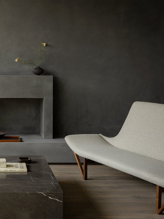 A light grey Pagode Sofa with wooden legs in a dark room, beside a Plinth Grand coffee table and a concrete fireplace.