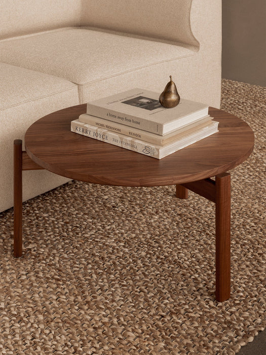 A round, walnut Passage Lounge Table on a woven rug beside an Eave Modular Sofa, styled with books and a paperweight.