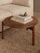 A round, walnut Passage Lounge Table on a woven rug beside an Eave Modular Sofa, styled with books and a paperweight.
