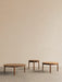 A set of three round, wooden Passage Lounge Tables in varying sizes, set against a plain beige wall on a grey floor.