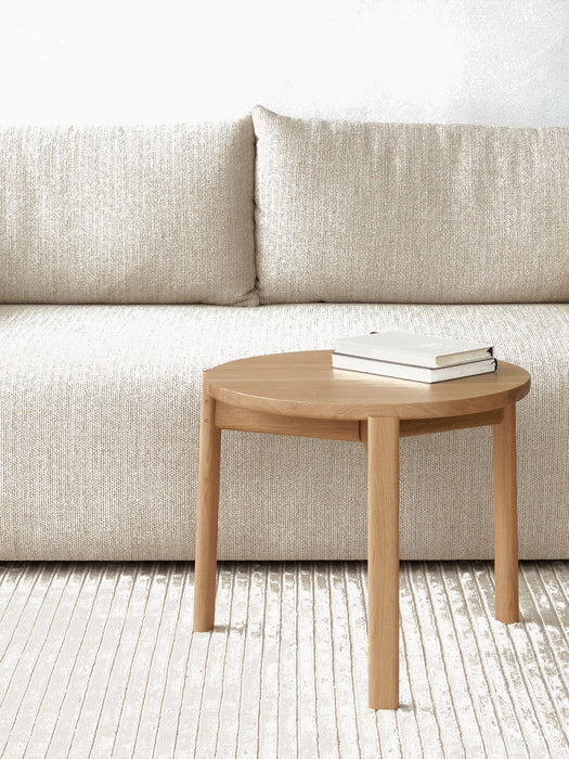 A close-up of a round oak Passage Lounge Table with two white books, set against a beige Offset Sofa and on a Houkime Rug.