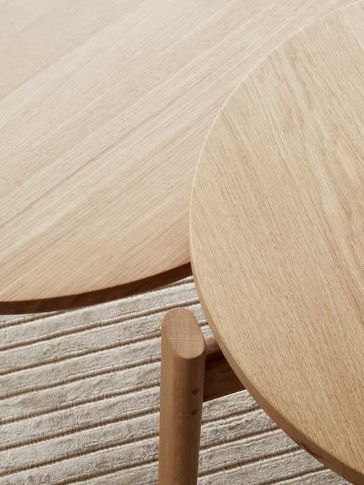 A close-up detail shot of the natural oak Passage Lounge Table's round top and leg, resting on the textured Houkime Rug.