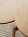 A close-up detail shot of the natural oak Passage Lounge Table's round top and leg, resting on the textured Houkime Rug.