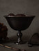 A still life image of the dark bronzed Pavot Bowl on a pedestal, filled with dark plums, set against a dark grey background.