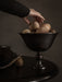 A hand in a dark sweater reaches for a stone inside the dark bronze Pavot Bowl, which sits on a table in a moody setting.
