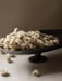 A close-up shot of the Pavot Centerpiece, a dark pedestal bowl, overflowing with a mound of dried, off-white flowers.