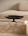 The dark bronze Pavot Centerpiece rests on a marble Plinth Low coffee table with a cream Eave sofa blurred in the background.
