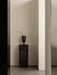 A tall Plinth Pedestal in black marble stands in a minimalist setting. The dark Pavot Vase holds two white calla lilies.