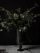 The dark metal Pavot Vase styled with a large bouquet of pale green flowers and foliage on a marble tabletop.
