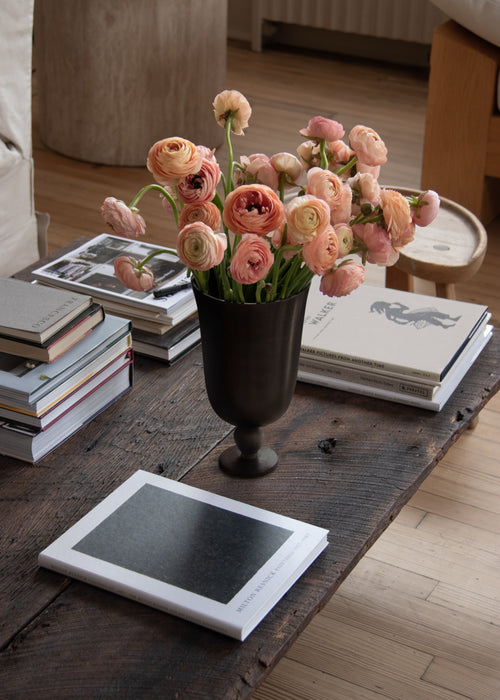 The Audo Pavot Vase, a dark chalice-shaped vessel, holds pink ranunculus flowers on a rustic wooden coffee table with books.