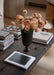 The Audo Pavot Vase, a dark chalice-shaped vessel, holds pink ranunculus flowers on a rustic wooden coffee table with books.