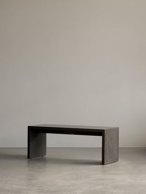 The dark marble Plinth Bridge, a long minimalist table, in a simple room with a concrete floor and grey wall.
