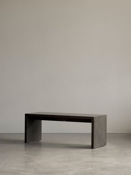 The dark marble Plinth Bridge, a long minimalist table, in a simple room with a concrete floor and grey wall.