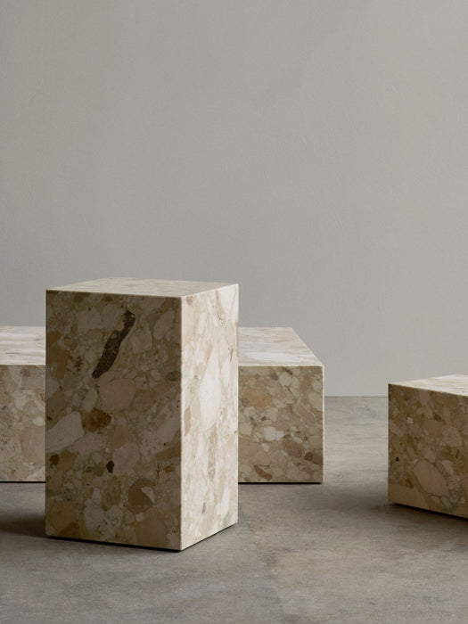 A collection of Plinth Low, Tall, and Grand tables in Kunis Breccia stone, arranged on a concrete floor against a pale wall.