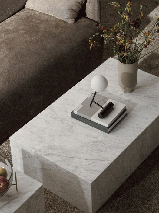 High-angle view of a white marble Plinth Low table styled with a Phare Lamp and a vase of flowers, next to a brown sofa.
