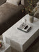 High-angle view of a white marble Plinth Low table styled with a Phare Lamp and a vase of flowers, next to a brown sofa.