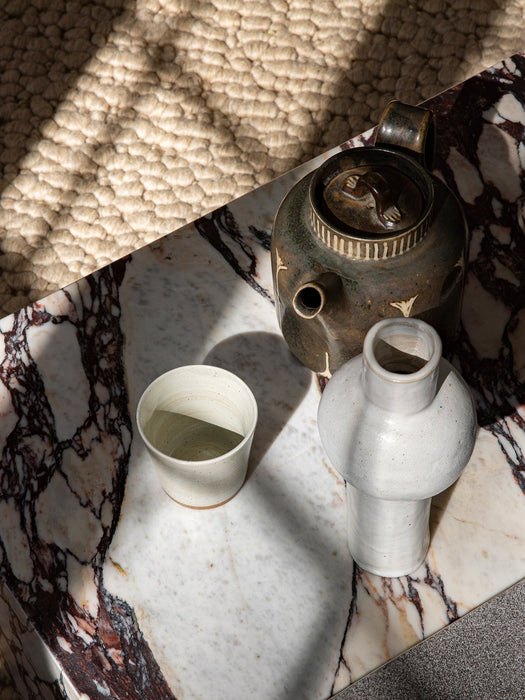 Close-up of the Plinth Low in Calacatta Viola marble, styled with a rustic ceramic teapot, cup, and vase under sunlight.