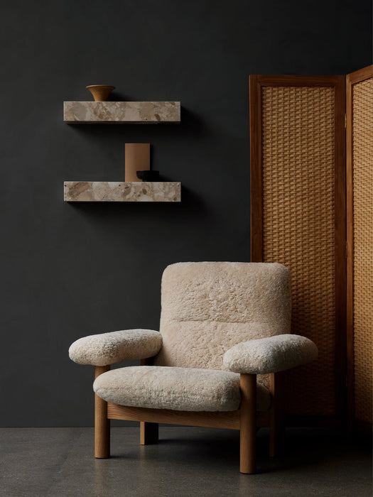 A cream sheepskin Brasilia Lounge Chair in a dark room with two marble Plinth Shelves on the wall and a rattan screen.