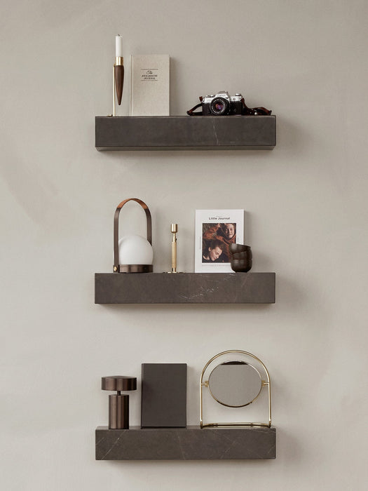 Three dark marble Plinth Shelves on a plain wall, styled with an Umanoff Candle Holder, lamps, a camera, and other decor.