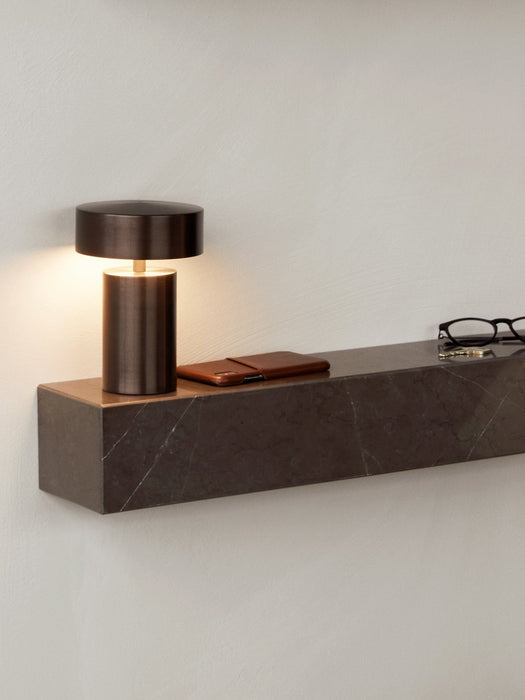 A close-up of the dark marble Plinth Shelf mounted on a wall, with a lit bronze Column Table Lamp, phone, and glasses on top.