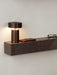 A close-up of the dark marble Plinth Shelf mounted on a wall, with a lit bronze Column Table Lamp, phone, and glasses on top.