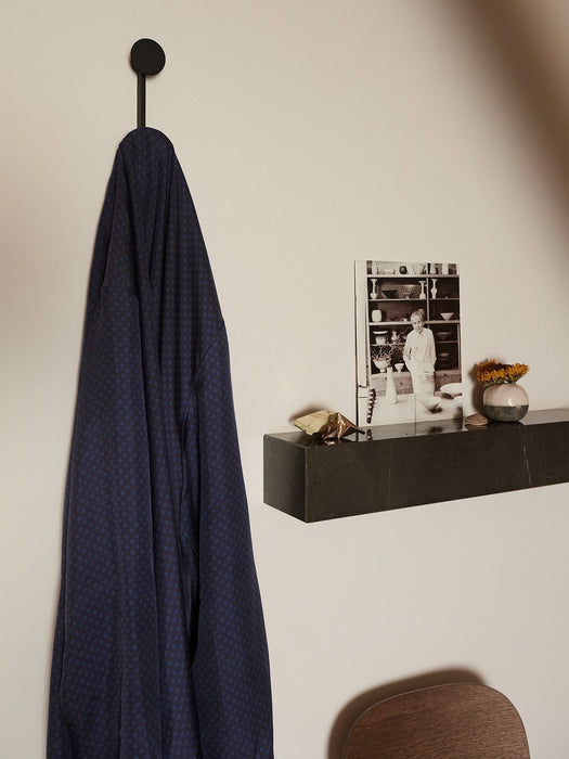 A black Afteroom Coat Hanger with a blue robe, next to a black marble Plinth Shelf holding a photo and a vase with flowers.