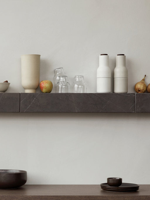A dark marble Plinth Shelf holds Bottle Grinders, a Cyclades Vase and fruit. Below, New Norm Dinnerware sits on a table.