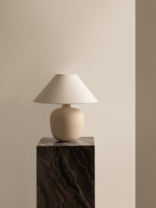 The Torso Table Lamp, with a beige ceramic base and linen shade, is placed on a tall Plinth made from Grey Galaxy marble.