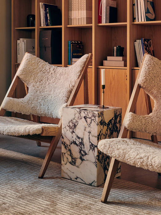 Two wooden Knitting Chairs with off-white sheepskin upholstery flank a marble Plinth side table in a cosy, book-lined room.
