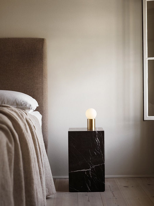 The Socket Lamp with its brass base and glowing orb bulb rests on a black marble Plinth next to a bed with a beige blanket.