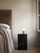 The Socket Lamp with its brass base and glowing orb bulb rests on a black marble Plinth next to a bed with a beige blanket.