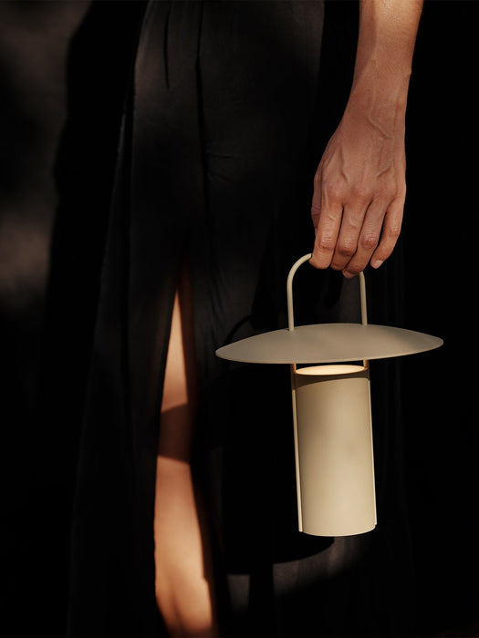 A person in a black dress holds the lit Ray Table Lamp in matte dusty green by its handle against a dark, moody background.