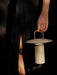 A person in a black dress holds the lit Ray Table Lamp in matte dusty green by its handle against a dark, moody background.