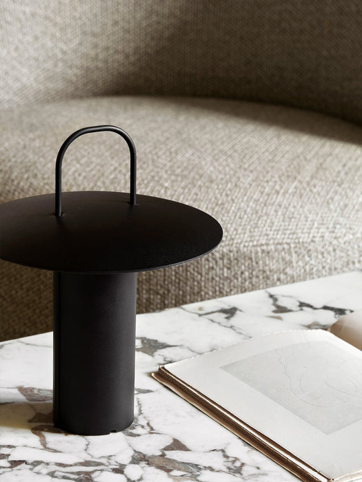 The black Ray Table Lamp, Portable, with its minimalist handle, sits on a white and grey marble table next to an open book.