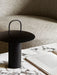 The black Ray Table Lamp, Portable, with its minimalist handle, sits on a white and grey marble table next to an open book.