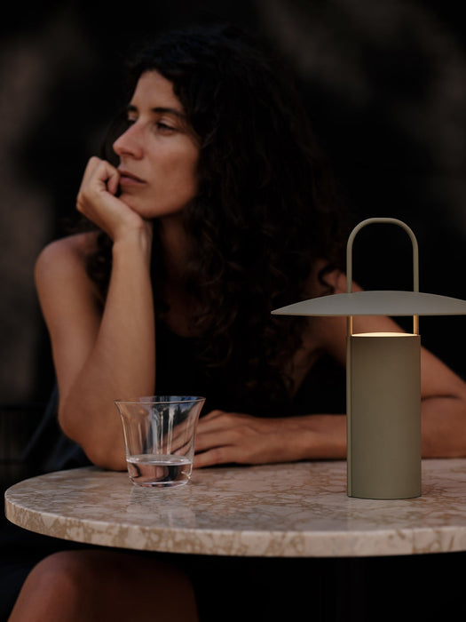 The Ray Table Lamp in matte dusty green, glowing on a marble Harbour Column Table next to a woman and a Strandgade glass.