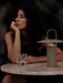 The Ray Table Lamp in matte dusty green, glowing on a marble Harbour Column Table next to a woman and a Strandgade glass.