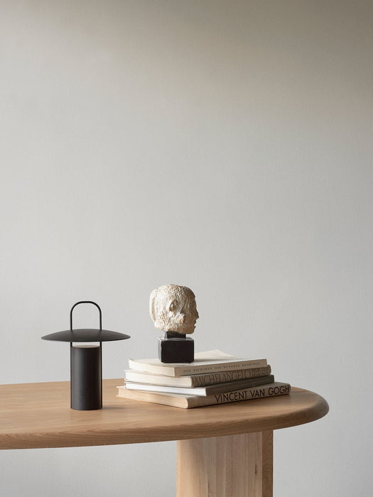 The black Ray Table Lamp, Portable, styled on a light oak table next to a stack of books with a small sculpture on top.