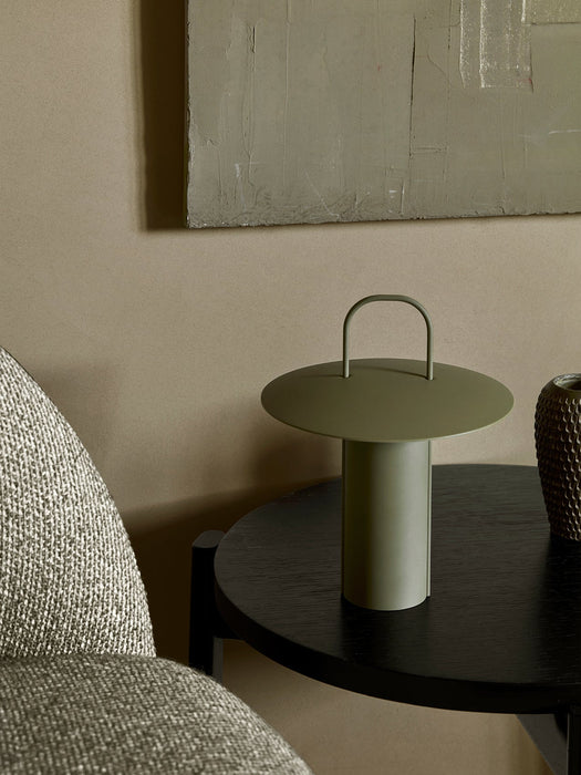 A close-up of the Ray Table Lamp in Matte Dusty Green, placed on a dark wooden side table next to a textured grey chair.