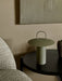 A close-up of the Ray Table Lamp in Matte Dusty Green, placed on a dark wooden side table next to a textured grey chair.