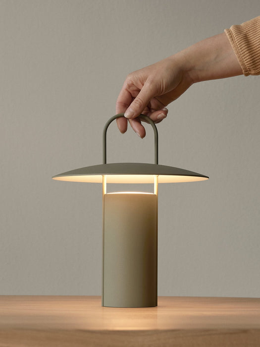 A hand holds the handle of the Matte Dusty Green Ray Table Lamp, Portable, which is switched on and sits on a wooden table.