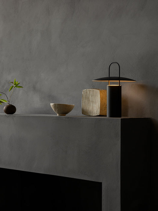 The black Ray Table Lamp provides a warm glow on a dark grey mantelpiece, styled with a ceramic bowl, a vase, and a sprig.