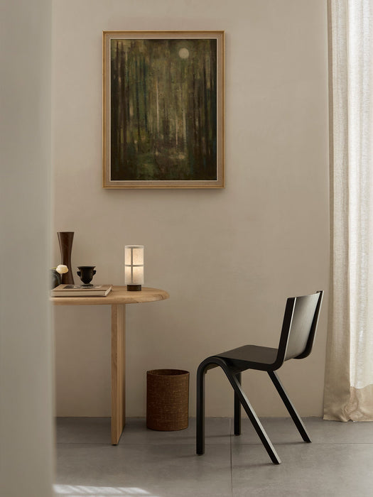 A black Ready Dining Chair stands beside a round wooden table. On the table is a lit Hashira Table Lamp, a book, and vases.